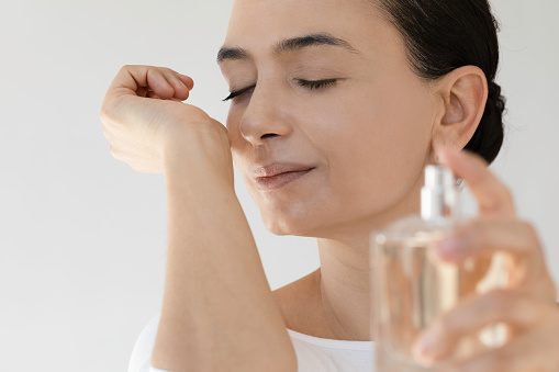 Unrecognizable woman sprays herself perfume on her wrist and is doing smelling gesture in front of white background.