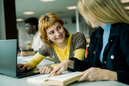 Positive young female student sitting at table with laptop in library and smiling at unrecognizable friend showing line in opened book while speaking and looking at each other