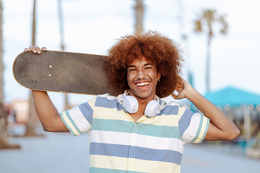 Portrait of a cheerful Black man with wireless headphones carrying a skateboard on his shoulders