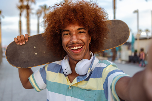 Portrait of a cheerful Black man with wireless headphones carrying a skateboard on his shoulder and looking at the camera