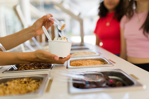 Close up of woman hands holding delicious ice cream or frozen yogurt at the toppings bar and selling to teen girls customers