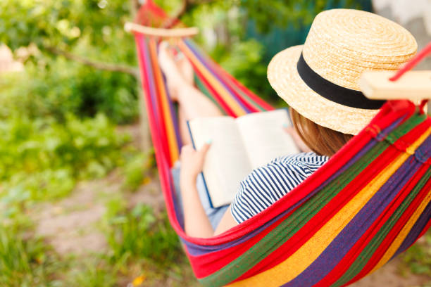 istock Young woman reading book while lying in comfortable hammock at green garden 1496737377