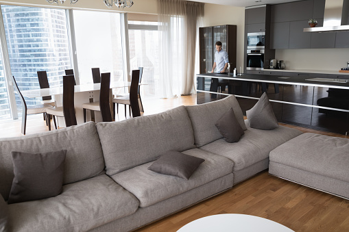 Elegant interior of modern urban apartment with large window and city skyscraper view. Hotel senior suite room, studio with big comfortable couch and young man in kitchen in background
