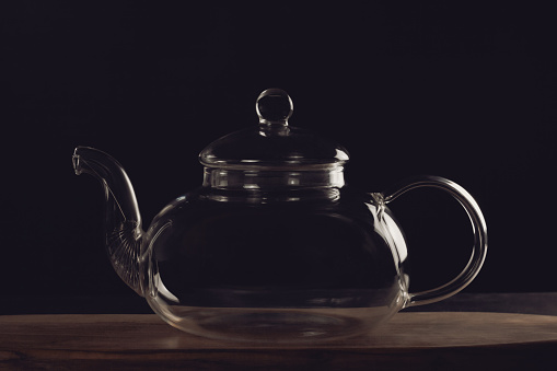 A beautiful glass teapot for brewing tea stands on a wooden board on a dark background. The empty teapot is transparent
