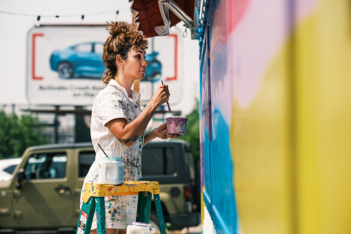 Mid adult Iranian woman artist painting  mural on shipping container, being converted in public food bank kiosk.  She is dressed in casual work clothes. Exterior of public sidewalk in  large North American City.