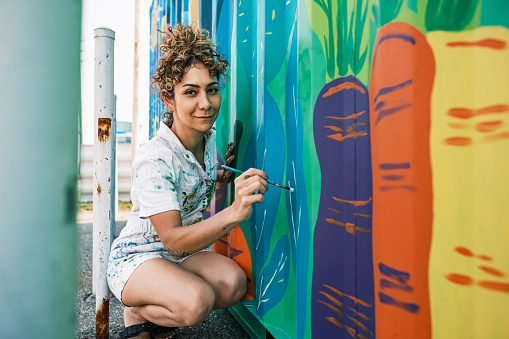 Mid adult Iranian woman artist painting  mural on shipping container, being converted in public food bank kiosk.  She is dressed in casual work clothes. Exterior of public sidewalk in  large North American City.