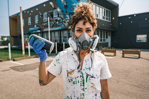 Mid adult Iranian woman artist painting  mural on shipping container, being converted in public food bank kiosk.  She is dressed in casual work clothes. Exterior of public sidewalk in  large North American City.