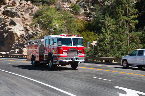 A red firetruck with emergency lights flashing passing cars as it races down a mountain road.