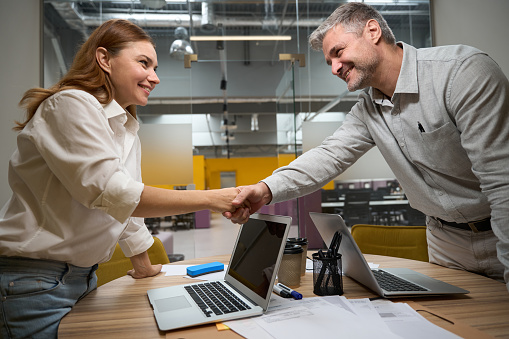 Young woman and man standing near the table with laptop and looking at each other, shaking hands