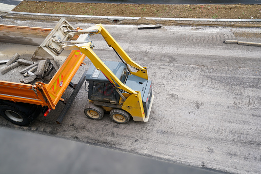 A small bulldozer loads the removed asphalt and road curbs into the dump tuck on road construction site. High angle view.