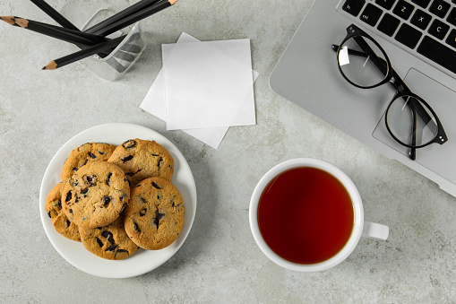Chocolate chip cookies, tea and laptop on light gray table at workplace, flat lay