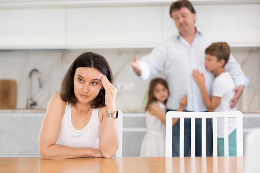 Offended mother sitting at the kitchen table, her husband, son and little daughter standing behind trying to dissuade her