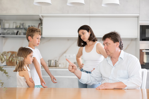 Angry parents scolding two children standing in front of them disappointedly in the kitchen