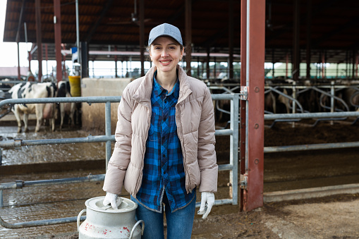 Portrait of young adult smiling woman farm worker holding big milk can at cowshed on dairy farm