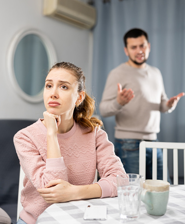 Young adult couple quarreling at home, woman sitting at table and ignoring arguing man