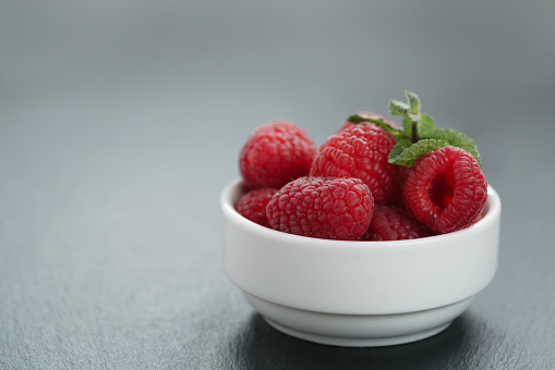 fresh raspberries with mint leaves in white bowl on slate board, shallow focus