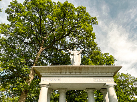 Jesus Figure Preaching atop an Ornate Stone Base with Columns. Set against a tall, leafy oak tree and a cloudy blue sky. Located within a cemetery in Springfield, Illinois, USA.