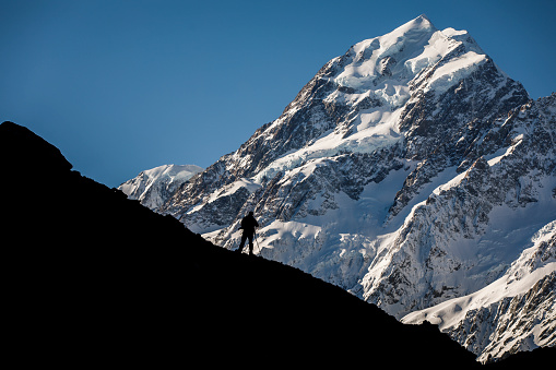 Nature scene of silhouetted man taking photos of large awe-inspiring snow covered mountain. Photographed in New Zealand.