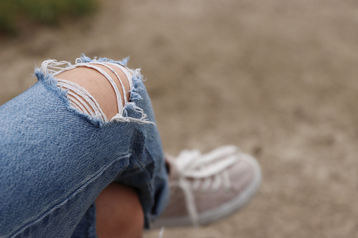 Young girl wearing ripped blue jeans with big hole on the knee is sitting on the bank crossed her legs. Woman in destroyed distressed denim. Close up of Textile texture. Selective focus, blurred
