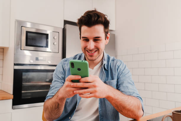 istock Young caucasian man playing a video game with a cellphone sitting at home kitchen. Happy guy using a smartphone app to send messages on social media. Teenage male smiling browsing with a mobile phone 1496767447