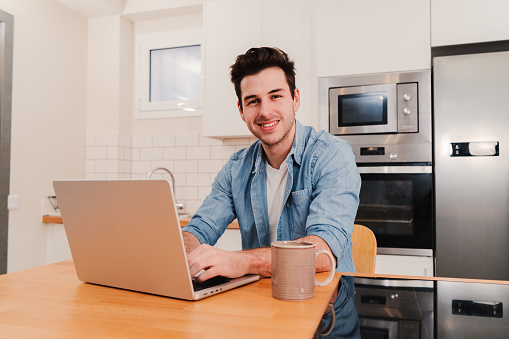 Handsome caucasian young man working remotely from home with a laptop looking at camera. Front view of happy male entrepreneur smiling using Internet wireless with a computer sitting at kitchen room. . High quality photo