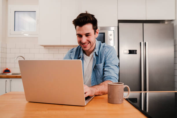 istock Handsome caucasian man working with a laptop computer at home kitchen drinking coffee and enjoying the morning. Happy young entrepreneur or student searching information on internet. Freelance work concept 1496767798