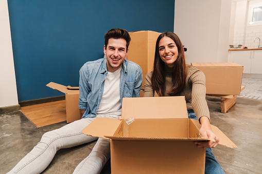 young caucasian couple smiling making a move to their new home, using cardboard boxes to carry their things. Husband and wife sitting on the floor of their apartment unpacking boxes. Loan mortgage concept. High quality photo