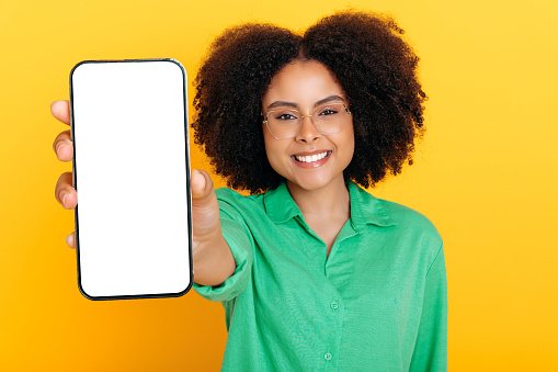 Trendy lovely brazilian or hispanic woman with glasses, in casual shirt, shows mobile phone with blank white mockup screen for presentation or advertising, smiles at camera, isolated yellow background