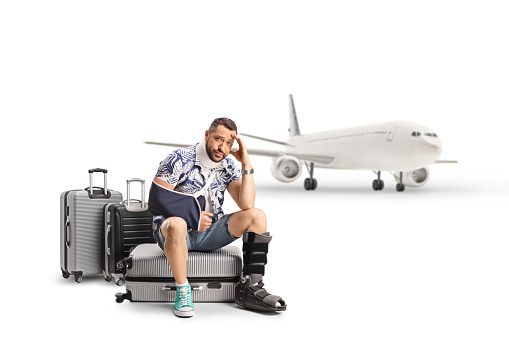 Sad male tourist with an injured arm and leg sitting on suitcases in front of an airplane isolated on white background