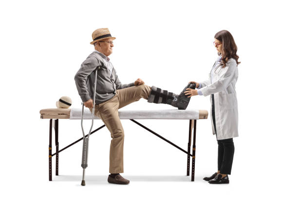 elderly man with a leg brace sitting on a bed and a female doctor checking his foot - physical injury men orthopedic equipment isolated on white imagens e fotografias de stock