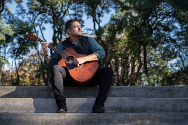 istock Guitarist sitting in a park playing his guitar 1496807438