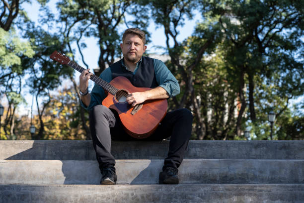 istock Guitarist sitting in a park playing his guitar 1496807439