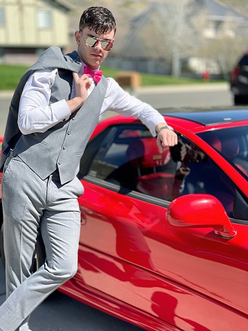 Handsome teenage boy, 17-18 years, grey tuxedo formalwear. Young adult, jacket over shoulder, legs crossed leaning on car looking cool feeling confident. High school memories, prom date.