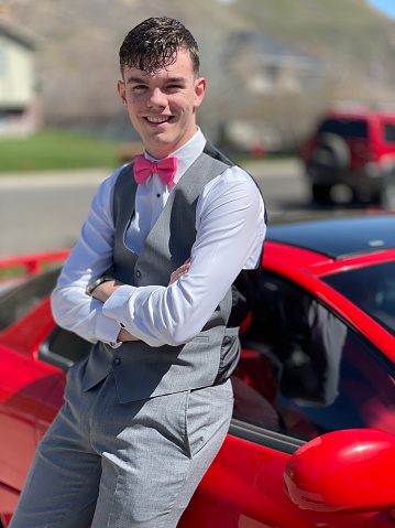 One teenage boy, smiling happy looking at camera, arms folded leaning on car. Confident young adult, gray tuxedo, ready for prom. High school student, memories, special occasion.