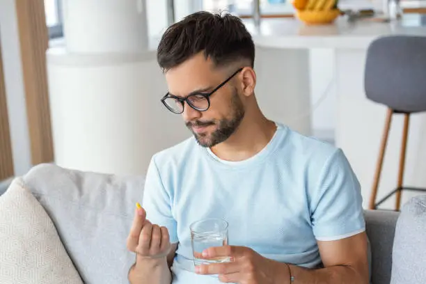 Photo of Young man hold pills and glass of water in his hands. Concept of healthcare and medicine, patient take daily dose of prescribed medicament, feel sick, antibiotics, painkillers or antidepressants. Close up