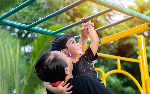 Happy Asian boy try to hang the green bar by his mother holding and helping him at out door playground under the big tree. Selective focus.