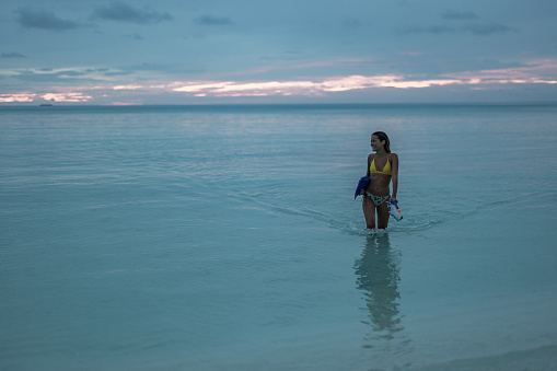 Young happy woman walking through sea with her diving equipment in the evening. Copy space.