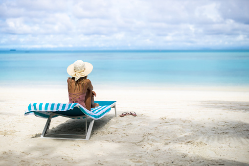 Rear view of a woman with sun hat sitting on a deck chair on the beach and looking at view. Copy space.