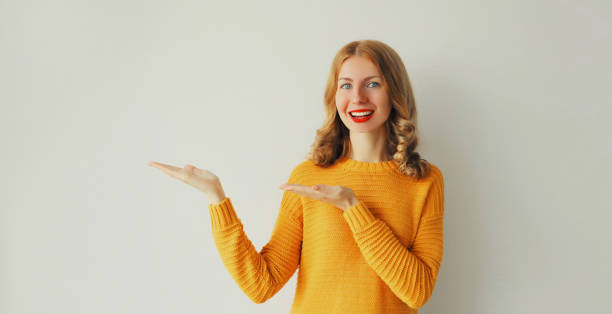 Portrait of happy smiling young woman holding something on her palm in hands pointing to the side wearing yellow knitted sweater on white background, blank copy space stock photo