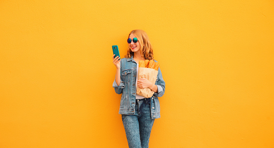 Portrait of happy smiling young woman holding phone grocery shopping paper bag with long white bread baguette on orange background