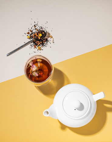 Creative flat lay of a glass cup of herbal tea with rose flowers on a yellow and white background with teapot and hard shadow. Food and drink concept. Top view and copy space.