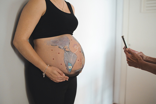 Tender scene of a multi-ethnic lesbian couple photographing the pregnant woman's belly drawing at home.