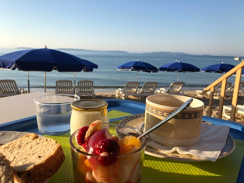 A table set with breakfast, including bread, fruits, and two mugs of coffee on the beach
