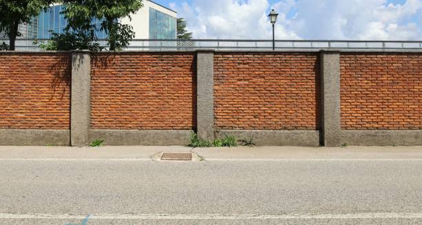 istock Surrounding brick wall with concrete columns with cloudy sky, street lamp and modern building on behind. Urban road in front. 1496858664