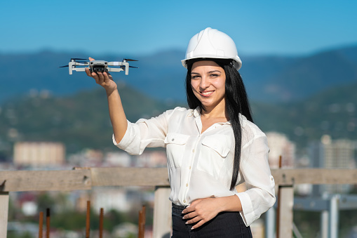 Smiling beautiful female geodesist engineer in a white helmet holds a drone at arm's length on a construction site and looks into the camera. Architectural Engineer and Safety Inspector