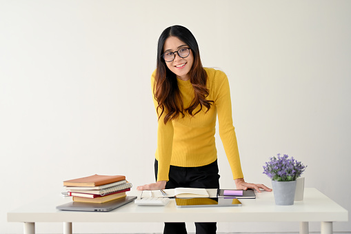 An attractive and happy young Asian female college student or office worker in glasses and casual clothes bends over at the table and smiles at the camera. isolated on a white background