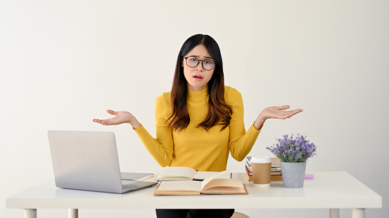 A puzzled and doubtful young Asian female college student or office worker in glasses sits at her desk, shrugging her shoulders with hesitation. don't know the answer, unsure and uncertain.