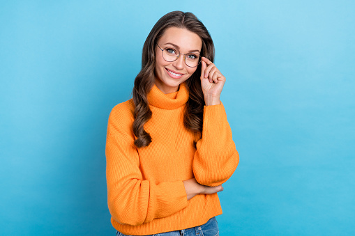 Photo of adorable smart woman wear orange sweater smiling arm spectacles isolated blue color background.
