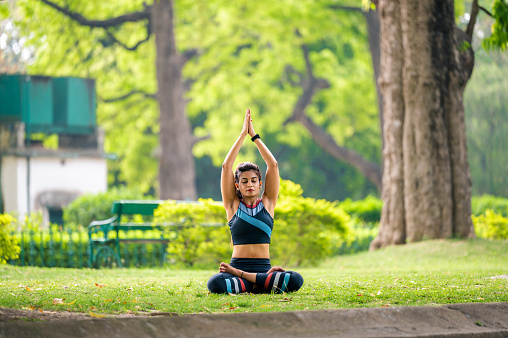 Young fitness woman sitting cross legged on grass in yoga posture, with arms raised over head in prayer pose and doing meditation at park in morning.