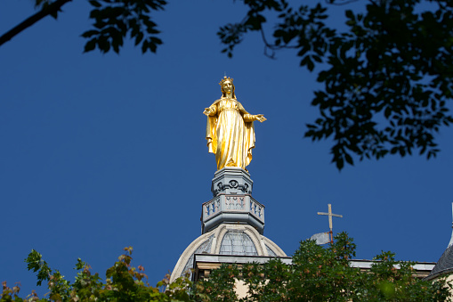 Lyon, Rhone Alpes, France \nMay 27, 2023\nPhoto of the gigantic gold statue of the holy virgin of the basilica Notre Dame de Fourvière on a large blue sky with trees in the foreground
